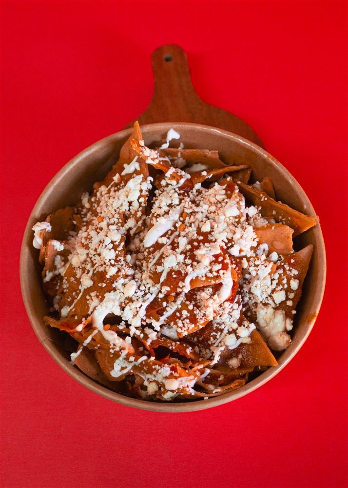 Chilaquiles rojos con queso y crema servidos en cazuela de barro sobre tabla de madera y fondo rojo.