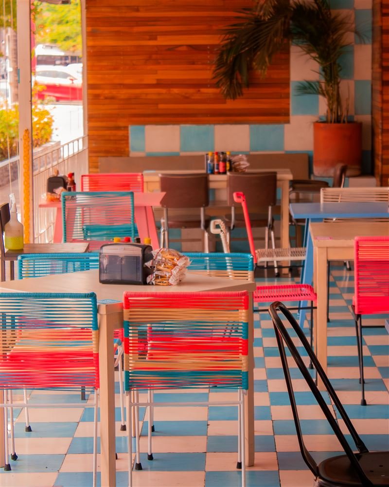Interior de local con sillas de acapulco en rojo y azul, piso de mosaico y muro de madera.