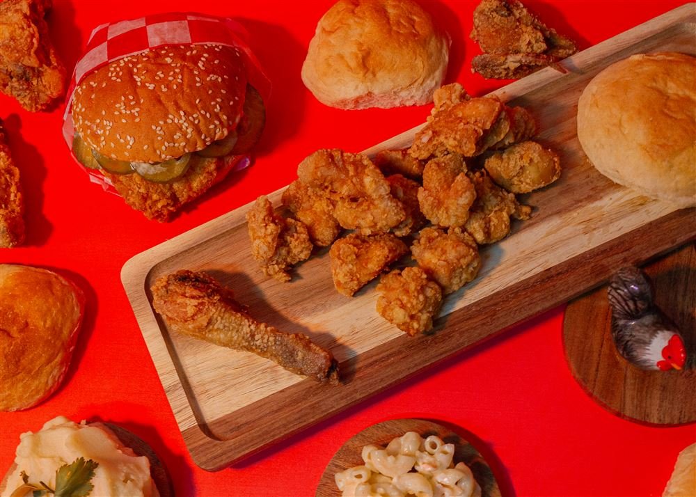 Tabla de madera con piezas de pollo frito y boneless, rodeada de hamburguesas y guarniciones sobre fondo rojo.